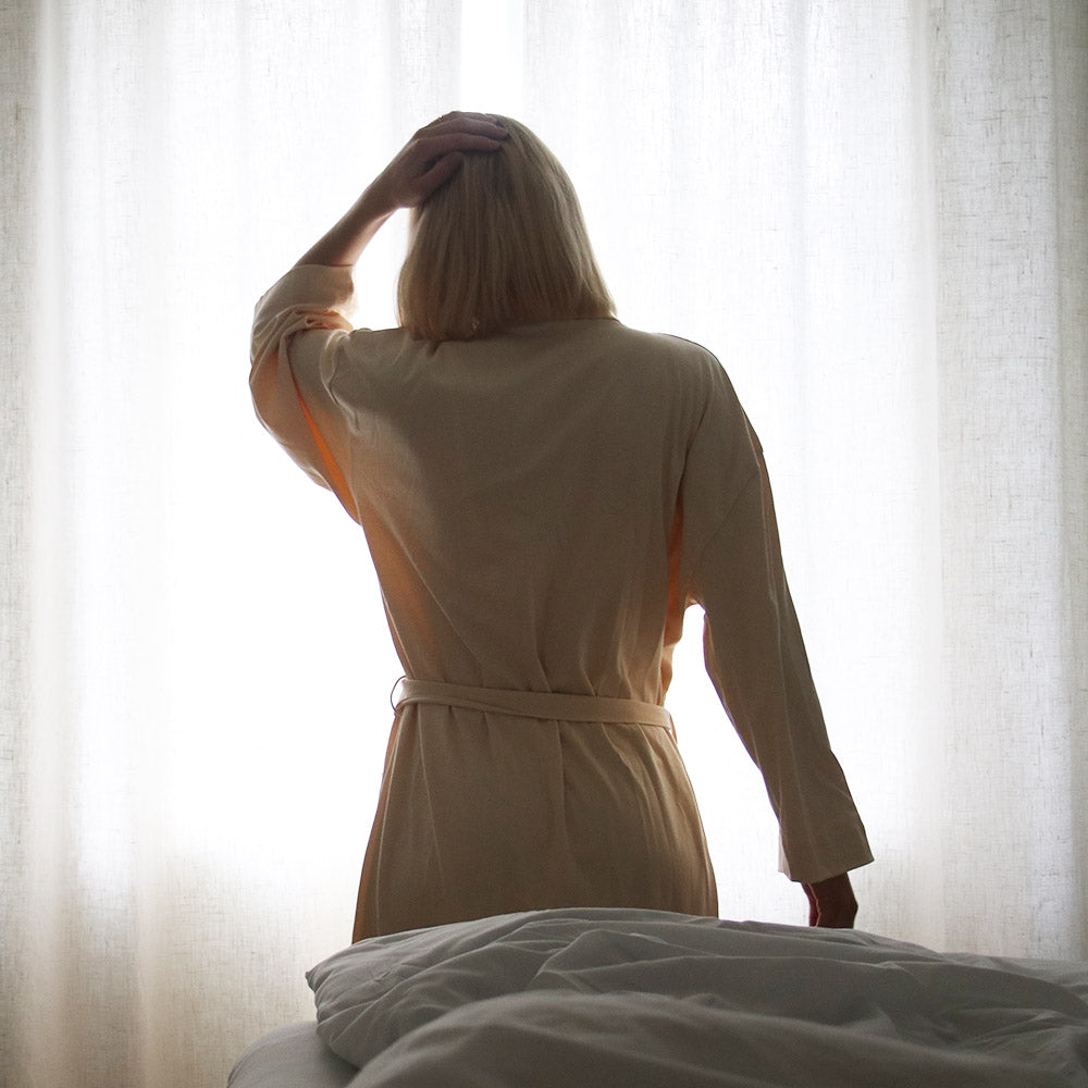 Woman facing a window wearing a beige morning robe in 100% organic pima cotton from The Sleepy Collection
